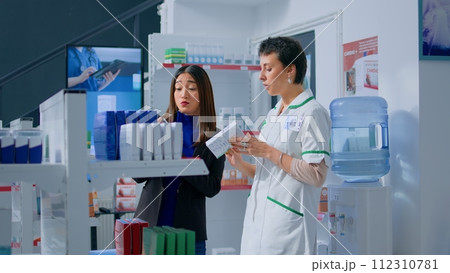 Shopper in chemist shop asking pharmacy employee about product information, trying to find right items on prescription list. Adept druggist helping customer choose fitting sickness medication Shopper in chemist shop asking pharmacy employee about product information, trying to find right items on prescription list. Adept druggist helping customer choose fitting sickness medication 112310781