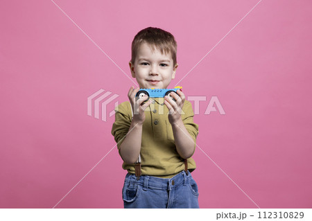 Small cute child playing around the studio with a blue car, having fun and recreating in front of camera. Cheerful smiling little boy holding a plastic vehicle to play and enjoy leisure activity. 112310829