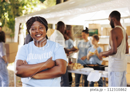 Enthusiastic black woman wearing blue t-shirt with arms crossed looks at camera. Portrait shot of african american female volunteer, ready to provide humanitarian aid to poor, needy and less fortunate Enthusiastic black woman wearing blue t-shirt with arms crossed looks at camera. Portrait shot of african american female volunteer, ready to provide humanitarian aid to poor, needy and less fortunate 112310862
