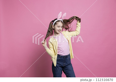 Smiling confident toddler dancing around in the studio with bunny ears, feeling happy and excited about easter event holiday. Young kid being joyful against pink background, wearing pigtails. 112310870
