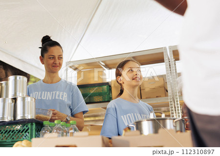 Caucasian mother and daughter volunteering at food drive, providing free food to the poor and needy. Female charity workers helping and giving essential items to homeless people and less fortunate. Caucasian mother and daughter volunteering at food drive, providing free food to the poor and needy. Female charity workers helping and giving essential items to homeless people and less fortunate. 112310897