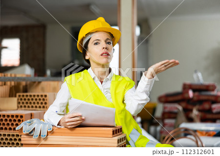 Woman foreman in a yellow vest and a protective helmet points with her hand somewhere in brick warehouse Woman foreman in a yellow vest and a protective helmet points with her hand somewhere in brick warehouse 112312601