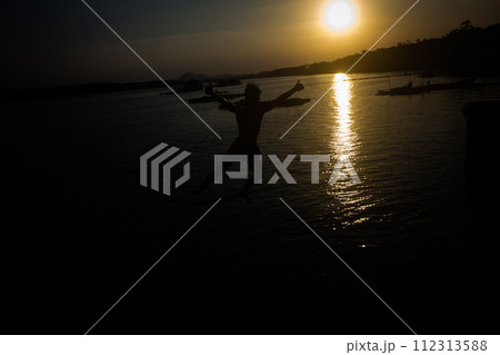 Silhouette of a man in shorts jumping into the water at a seaside pier. 112313588