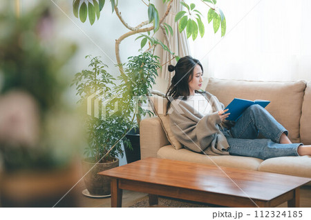 緑溢れるインテリアに囲まれてソファーに座りながら読書する女性 112324185