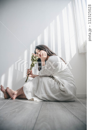 窓際で床に座り花を持つ白いワンピースの女性 112325028