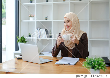 Asian Muslim business woman holding a coffee mug sitting in the office looking out the window. business people, diversity and office concept 112329003