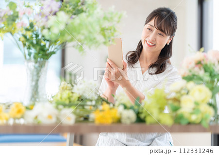 リビングで趣味のフラワーアレンジメントをスマホで撮影するミドルの女性 112331246