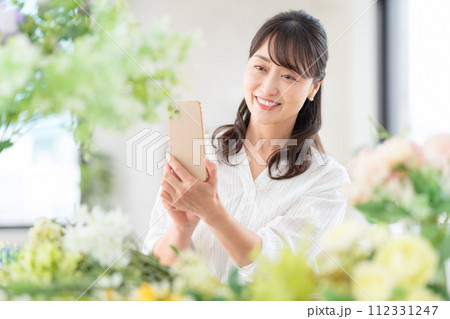 リビングで趣味のフラワーアレンジメントをスマホで撮影するミドルの女性 112331247