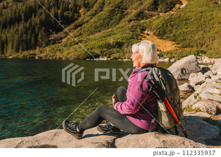 Rear shot of traveler senior woman in sportswear sitting on the rocky shore and making selfie on smart phone or video. Taking photo of green hills and mountains on Morskie Oko lake. Hiking travel and 112335937