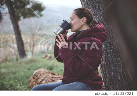 Happy relaxed young woman drinking hot tea from a thermos mug in the early spring forest Happy relaxed young woman drinking hot tea from a thermos mug in the early spring forest 112337021