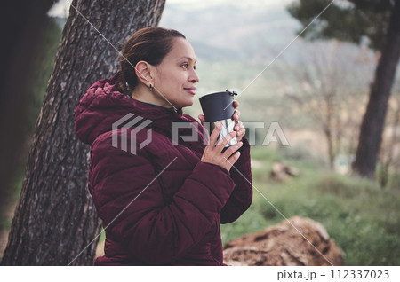 Young woman holding a thermos mug, enjoying spring nature in the forest, smiling and dreamily looking into the distance 112337023