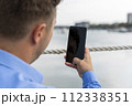 Man holding smartphone in his hand beside the river and making photographs 112338351