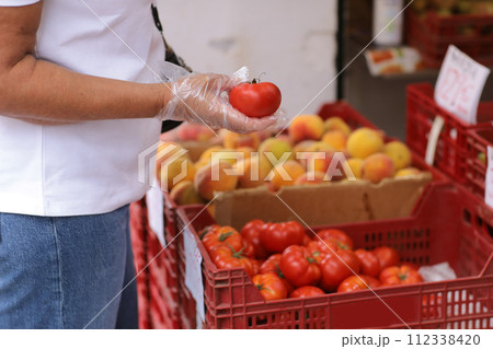 Unrecognisable overweight woman in disposable plastic gloves chooses fresh red tomatoes, buys vegetables in a grocery store or supermarket. Concept of shopping groceries, healthy lifestyle, hygiene. Unrecognisable overweight woman in disposable plastic gloves chooses fresh red tomatoes, buys vegetables in a grocery store or supermarket. Concept of shopping groceries, healthy lifestyle, hygiene. 112338420