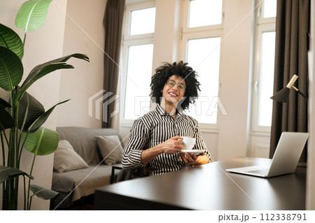 Smiling young man in striped shirt sitting at the table and having coffee Smiling young man in striped shirt sitting at the table and having coffee 112338791