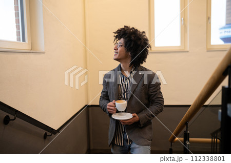 Curly-haired young man in eyeglasses at the stairs with a cup in hands 112338801