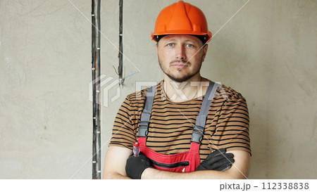 Renovation apartment. Portrait of mature confident male foreman constructor repairman with crossed hands in safety hard hat and overalls looks at camera, holds screwdriver. Empty concrete walls. Renovation apartment. Portrait of mature confident male foreman constructor repairman with crossed hands in safety hard hat and overalls looks at camera, holds screwdriver. Empty concrete walls. 112338838
