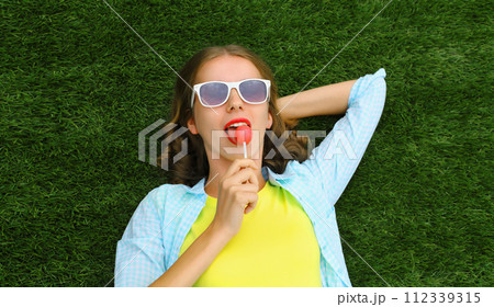 Happy teenager girl resting with lollipop lying on green grass in summer park 112339315