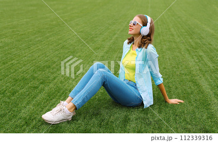 Happy smiling young woman listening to music in headphones while lying on grass in summer park 112339316
