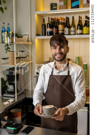 Man wearing apron holding coffee cup at cafe counter ensuring efficient service for customers Man wearing apron holding coffee cup at cafe counter ensuring efficient service for customers 112339400