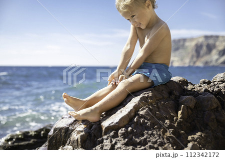Cheerful smiling little blond boy in blue swimming trunk sitting stone against backdrop sea  112342172