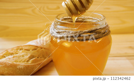 Golden fresh honey drips from a wooden spoon into a jar. There is a baguette nearby. Healthy eating. Golden fresh honey drips from a wooden spoon into a jar. There is a baguette nearby. Healthy eating. 112342462