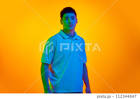 Young Asian man with calm facial expression looking at camera in vibrant neon light against gradient orange background. Young Asian man with calm facial expression looking at camera in vibrant neon light against gradient orange background. 112344847
