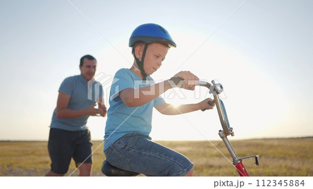 family play in the park. father teaching son to ride a bike. happy family kid dream concept. son learn to ride a bike silhouette. father supporting child son sunset riding bike summer in the park family play in the park. father teaching son to ride a bike. happy family kid dream concept. son learn to ride a bike silhouette. father supporting child son sunset riding bike summer in the park 112345884