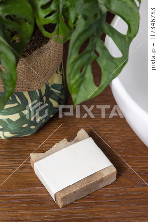Soap bar with blank label on bath wooden countertop near monstera plant close up, mockup 112346783