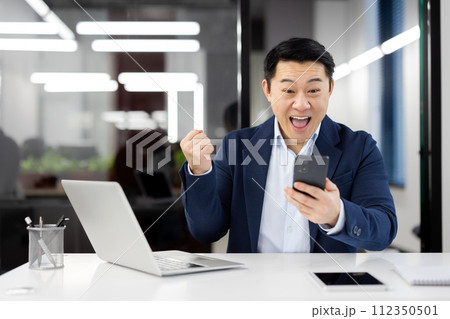 Joyful male entrepreneur in a suit punches the air in delight while looking at his phone in a well-lit office setup. 112350501
