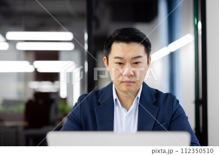Serious businessman working diligently at his laptop in a modern office setting, displaying focus and determination. Serious businessman working diligently at his laptop in a modern office setting, displaying focus and determination. 112350510