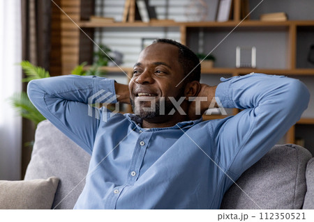 A content African American male sitting on a sofa at home, hands behind head, with a look of peaceful relaxation. 112350521