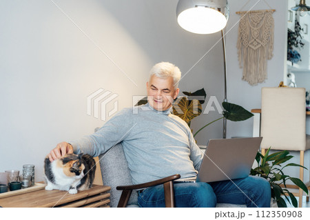 Relaxed gray hair middle-aged man stroking cat pet while working on laptop sitting in armchair at home. Cozy workspace in modern interior. Remote work at home. Senior entrepreneur manage business 112350908