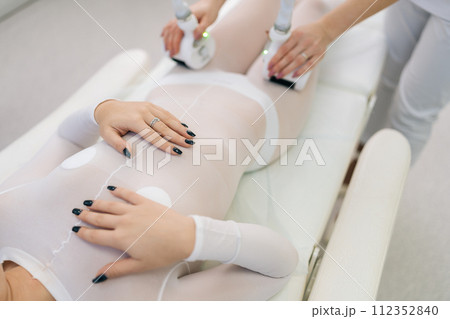 Top view of female client wearing special white suit receiving anti-cellulite massage in beauty salon. Closeup doctor hands manipulating hardware cosmetology LPG gadget for lifting body in spa center. 112352840