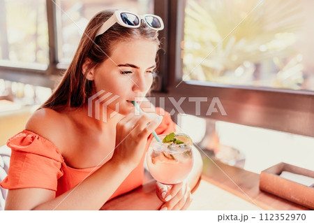 woman in a cafe seated, holding beverage. Dressed in an orange dress with her hair down. Bright interior, large windows allow natural light. Beverage consumption for refreshment. woman in a cafe seated, holding beverage. Dressed in an orange dress with her hair down. Bright interior, large windows allow natural light. Beverage consumption for refreshment. 112352970
