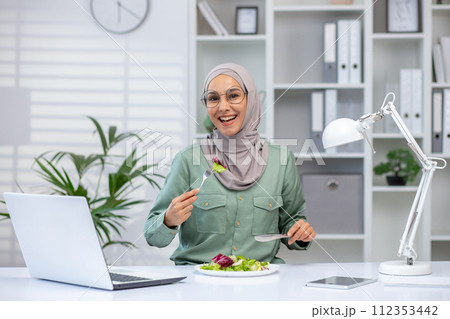 A joyful Muslim woman wearing a hijab eats a fresh salad at her desk, taking a break from work with her laptop and lamp beside. A joyful Muslim woman wearing a hijab eats a fresh salad at her desk, taking a break from work with her laptop and lamp beside. 112353442
