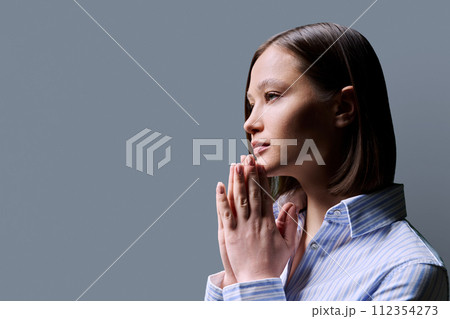 Close-up profile view of young woman with folded hands on gray background, copy space 112354273