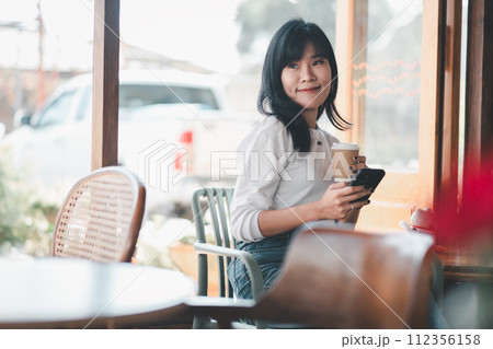 A relaxed young woman enjoys a moment to herself in a cafe, holding a coffee cup and browsing her smartphone, encapsulating a casual urban break. A relaxed young woman enjoys a moment to herself in a cafe, holding a coffee cup and browsing her smartphone, encapsulating a casual urban break. 112356158