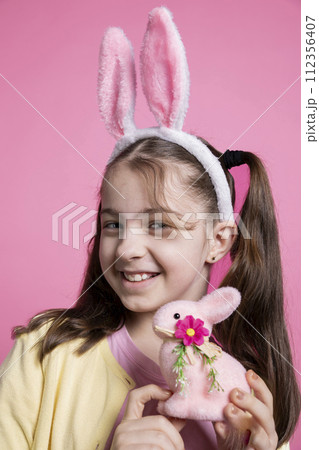 Sweet little girl posing with a cute pink stuffed rabbit on camera, showing her easter themed toys and decorations. Small child with bunny ears feeling happy and excited about celebration. Sweet little girl posing with a cute pink stuffed rabbit on camera, showing her easter themed toys and decorations. Small child with bunny ears feeling happy and excited about celebration. 112356407
