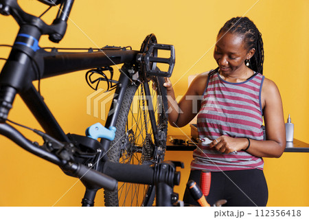 Energetic african american lady expertly repairs her bike wheel with specialized tools. Healthy committed black woman thoroughly inspecting and repairing bicycle tire against isolated background. 112356418