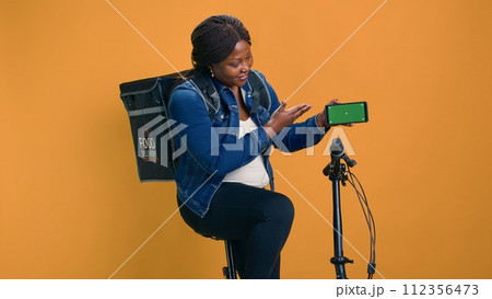 African american courier gripping cell phone showing green screen display while riding bicycle. Black woman demonstrates cutting-edge delivery app on mobile device with isolated mockup template. African american courier gripping cell phone showing green screen display while riding bicycle. Black woman demonstrates cutting-edge delivery app on mobile device with isolated mockup template. 112356473
