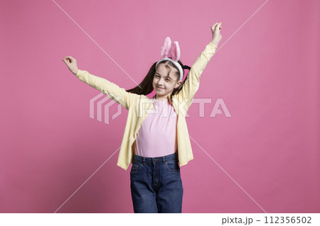 Sweet small child dancing in front of studio camera, posing with confidence and having fun. Little girl acting carefree with dance moves over pink background, wears bunny ears. Sweet small child dancing in front of studio camera, posing with confidence and having fun. Little girl acting carefree with dance moves over pink background, wears bunny ears. 112356502
