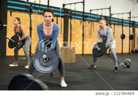 Muscular woman with group of sports people in activewear doing squats with barbell in gym fitness center 112360864