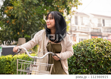 A beautiful young Asian woman in a cute dress walking on the footpath in the city with her bicycle. A beautiful young Asian woman in a cute dress walking on the footpath in the city with her bicycle. 112360915