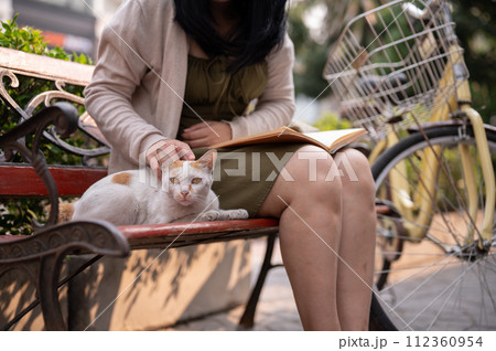 A beautiful Asian woman is playing with a stray cat while reading a book on a bench outdoors. 112360954