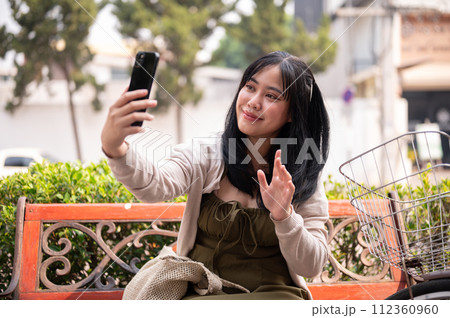 A positive Asian woman is talking on a video call with someone while relaxing on a bench outdoors. 112360960
