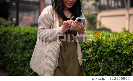 A happy Asian woman in casual clothes using her smartphone while walking in the city on a bright day A happy Asian woman in casual clothes using her smartphone while walking in the city on a bright day 112360996