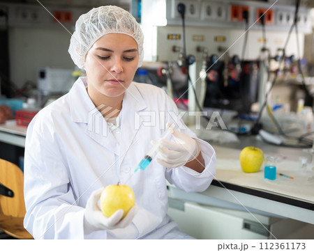 Young female scientist checking fruits for harmful elements, injecting syringe into apple in lab 112361173