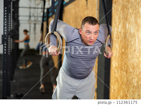 Adult male athlete with group working out on gymnastics rings at crossfit gym Adult male athlete with group working out on gymnastics rings at crossfit gym 112361175