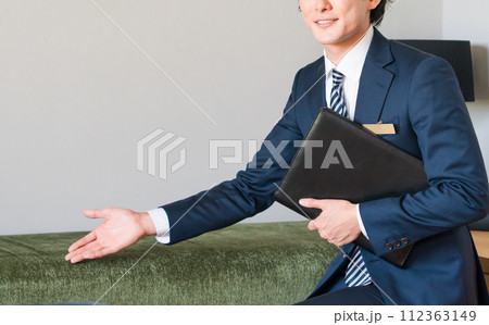モデルハウスのリビングでソファを案内・提案するスーツ姿のハウスメーカーの若い男性営業マン 112363149
