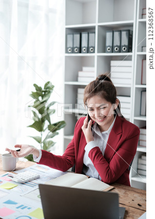 Happy Asian business women have the joy of talking on the smartphone, tablet, and laptop in the home office, Wearing a red suite. 112368567